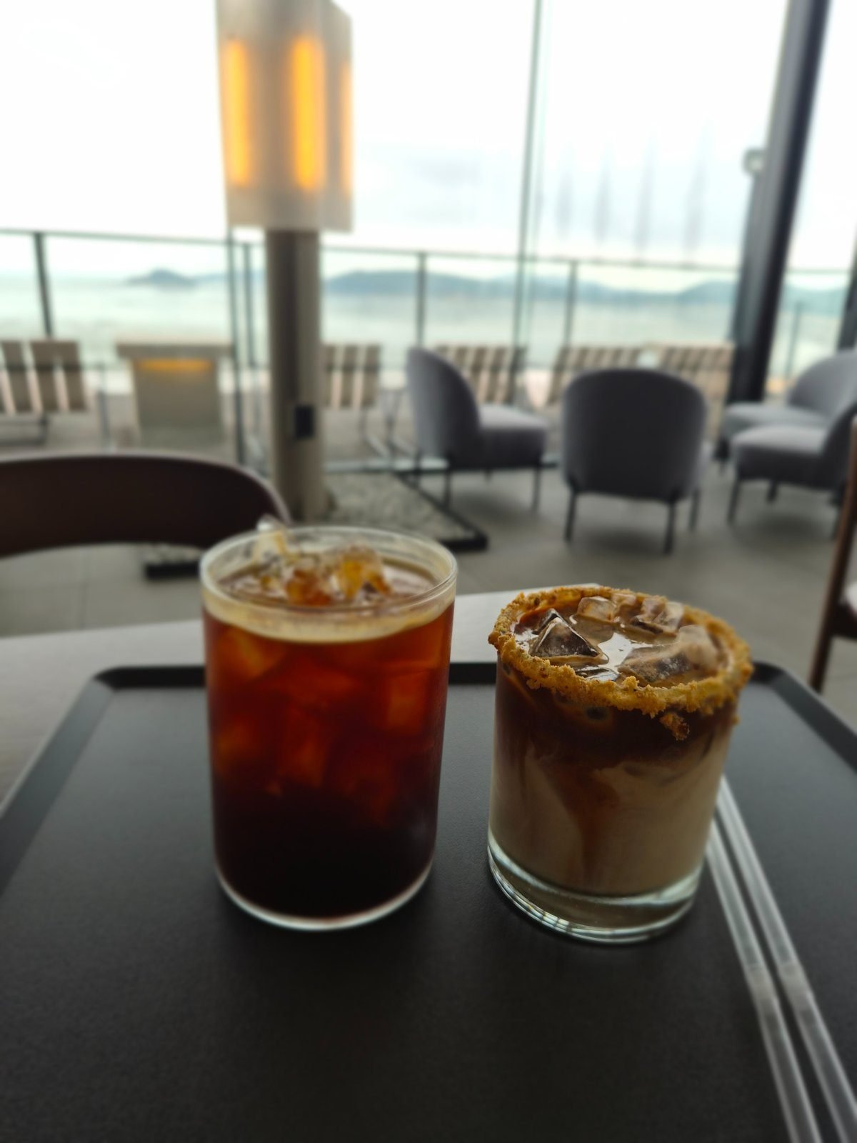 Iced americano and a nuts cream latte with hazelnut pieces on the rim, on a tray by the window at Emcliff cafe.