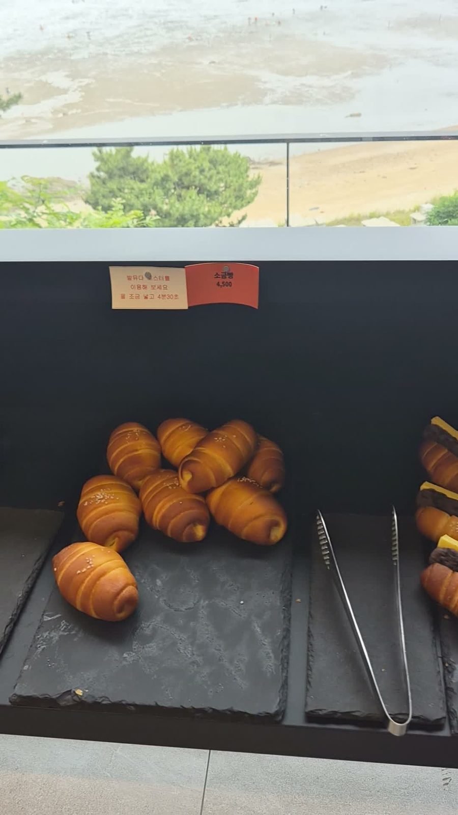 Cream-filled bread rolls on a slate tray at the Emcliff bakery counter, with Masian Beach visible through the window above.