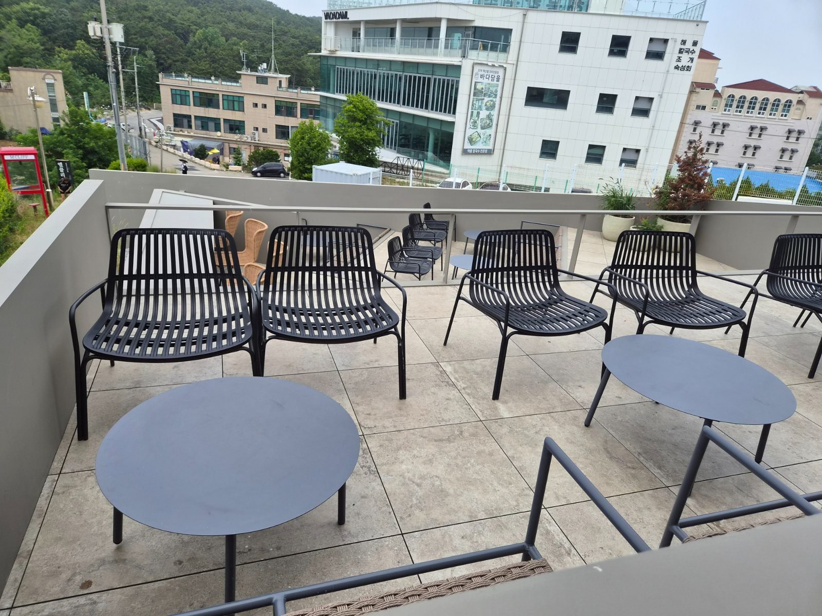 Outdoor terrace at Emcliff cafe with black rattan chairs and round tables, backed by the village and low wooded hills.
