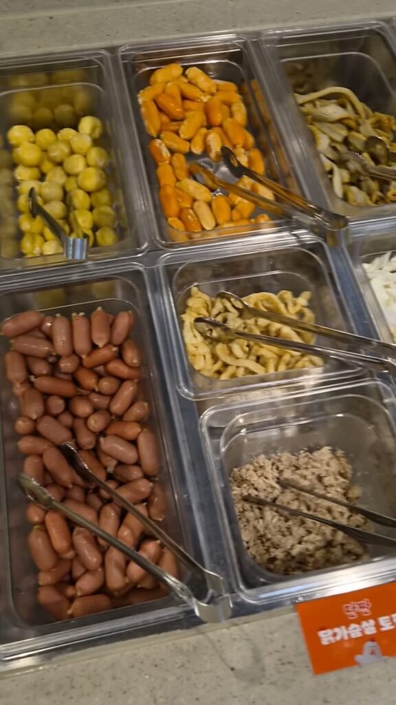 The self-serve ingredient bar at Dookki with trays of sweetcorn, fish cake, noodles, cocktail sausages and rice cakes
