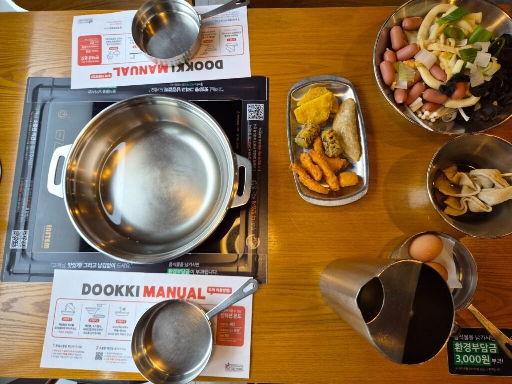 A table at Dookki with an induction hob, a steel pot, a printed DOOKKI MANUAL card and a green sticker warning of a 3,000 won charge for wasted food