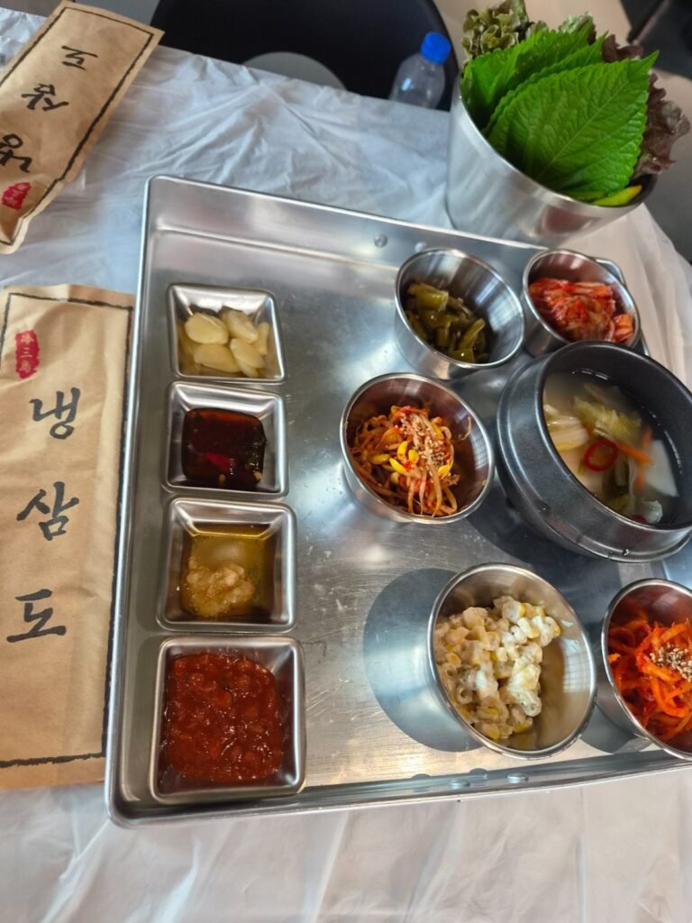 A steel tray of side dishes at Naengsamdo: garlic, ssamjang, chilli sauce, seasoned bean sprouts, radish salad, kimchi and sweetcorn