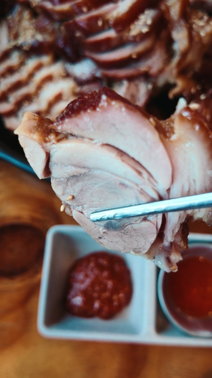 A slice of braised pork held on chopsticks above two dipping sauces