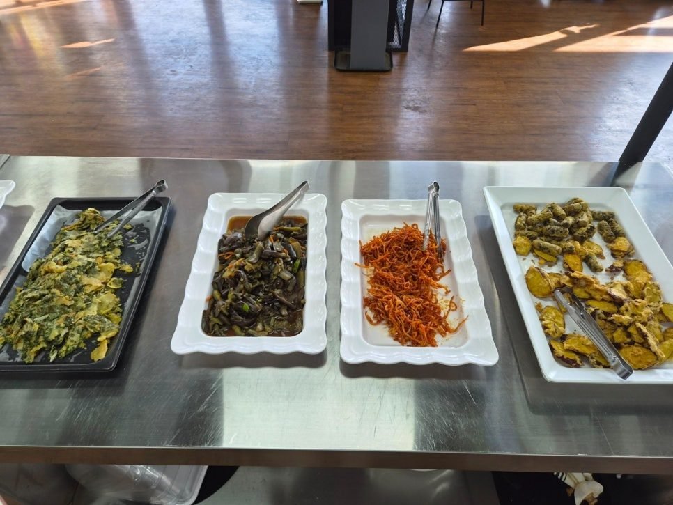 Steel counter with white rectangular trays of chive pancake, braised greens, seasoned dried squid and fritters, each with its own tongs