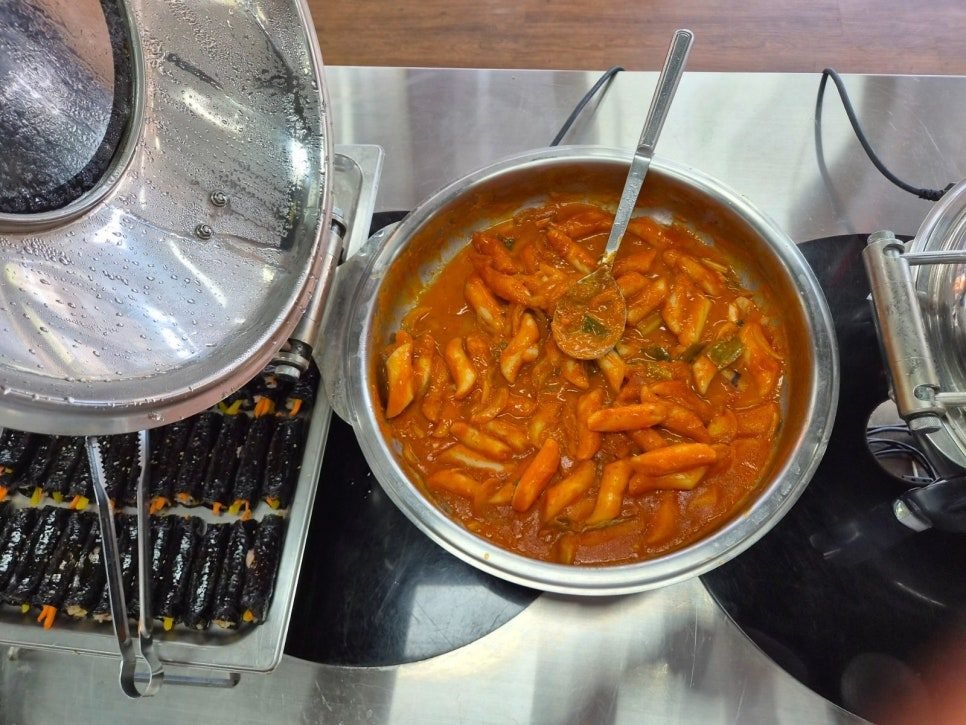 Steel pot of tteokbokki in red sauce beside rows of mini gimbap rolls on a serving counter