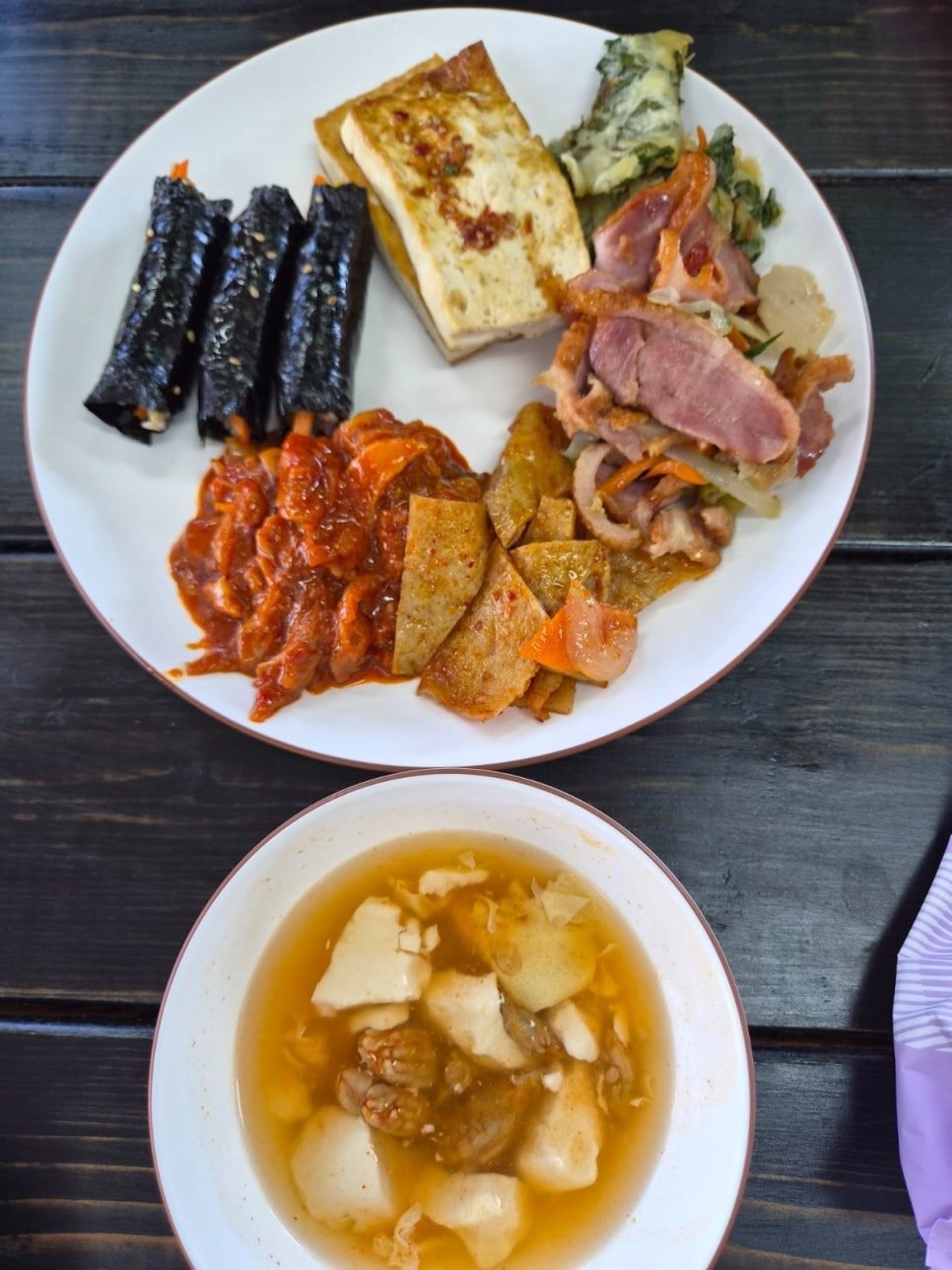White plate with mini gimbap, pan-fried tofu, smoked duck and fritters, next to a bowl of soft tofu stew on a dark wooden table