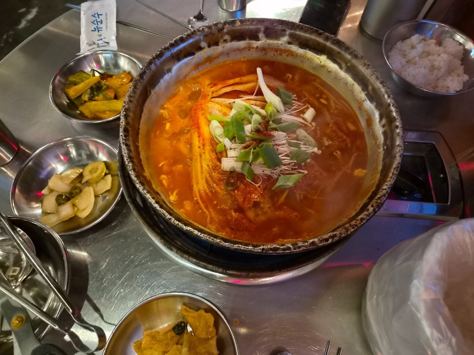 Kimchi stew in a wide hotpot on a table burner, with steel bowls of rice and side dishes around it