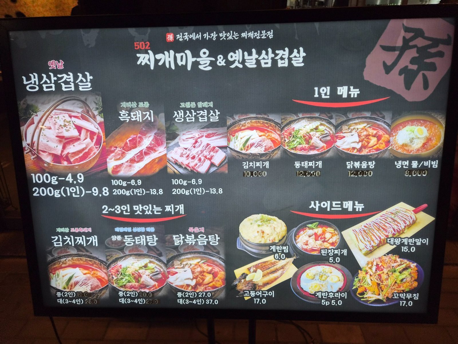 Backlit picture menu board at 502 Jjigae Maeul showing kimchi stew, pollack stew and chicken stew with prices