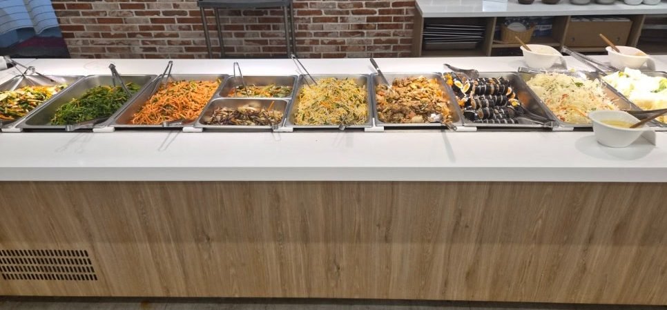 Eight steel trays on the buffet counter at Bangatgan holding greens, carrot salad, braised mushrooms, stir-fried vegetables, a braised meat dish, mini gimbap rolls and shredded cabbage salad.
