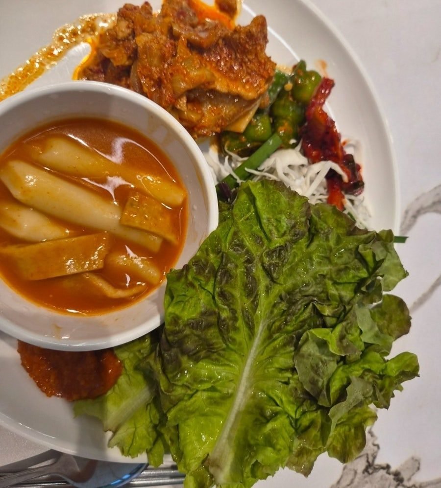 A plate holding spicy braised pork, a bowl of broth tteokbokki with flat fish cake, lettuce leaves, shredded cabbage and a green-chilli side dish.