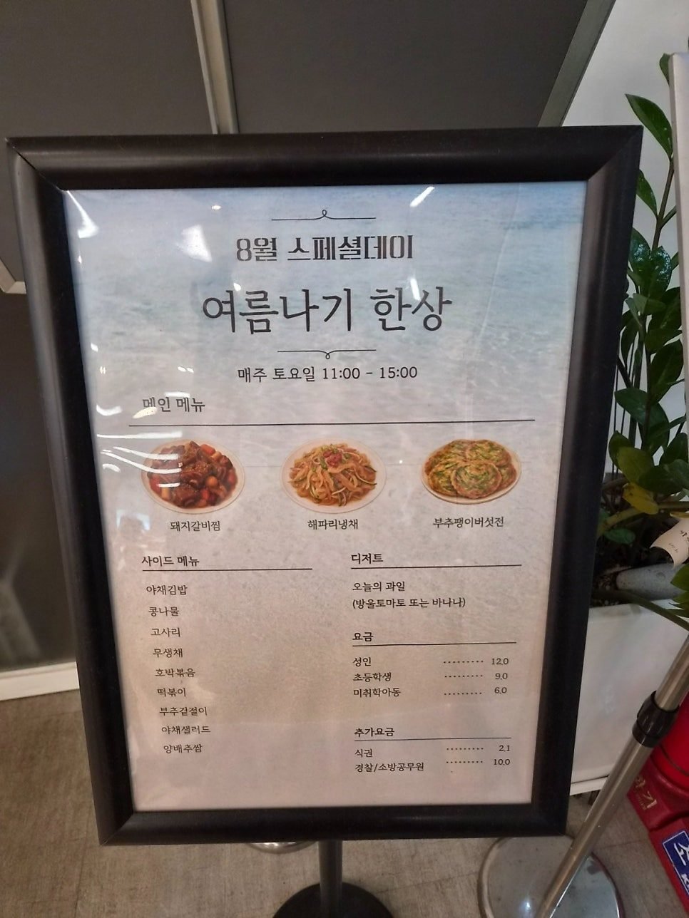 A framed poster headed 8월 스페셜데이 여름나기 한상, showing 돼지갈비찜, 해파리냉채 and 부추팽이버섯전, Saturdays 11:00 to 15:00, priced 12.0, 9.0 and 6.0.