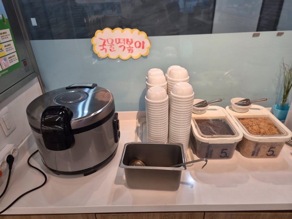 A self-serve counter with an electric warmer under a sign reading 국물떡볶이, stacks of white bowls, a ladle, and two lidded tubs of cereal.