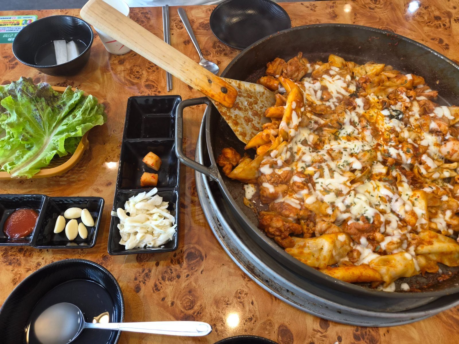 Cheese dak-galbi cooking on a round table griddle, chicken and rice cakes in red sauce under melted mozzarella, with a wooden spatula resting on the edge