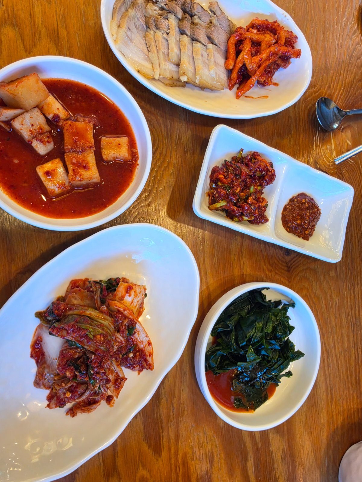 Side dishes on a wooden table: a plate of white boiled pork slices, red kimchi, spicy seasoned radish, blanched perilla greens and small dishes of salted seafood