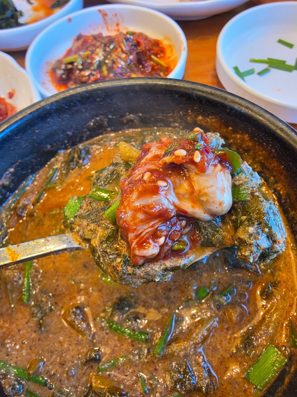 Close-up of chueotang broth with a spoonful of kimchi on top, green chilli rings and chopped chives at the edge of the pot