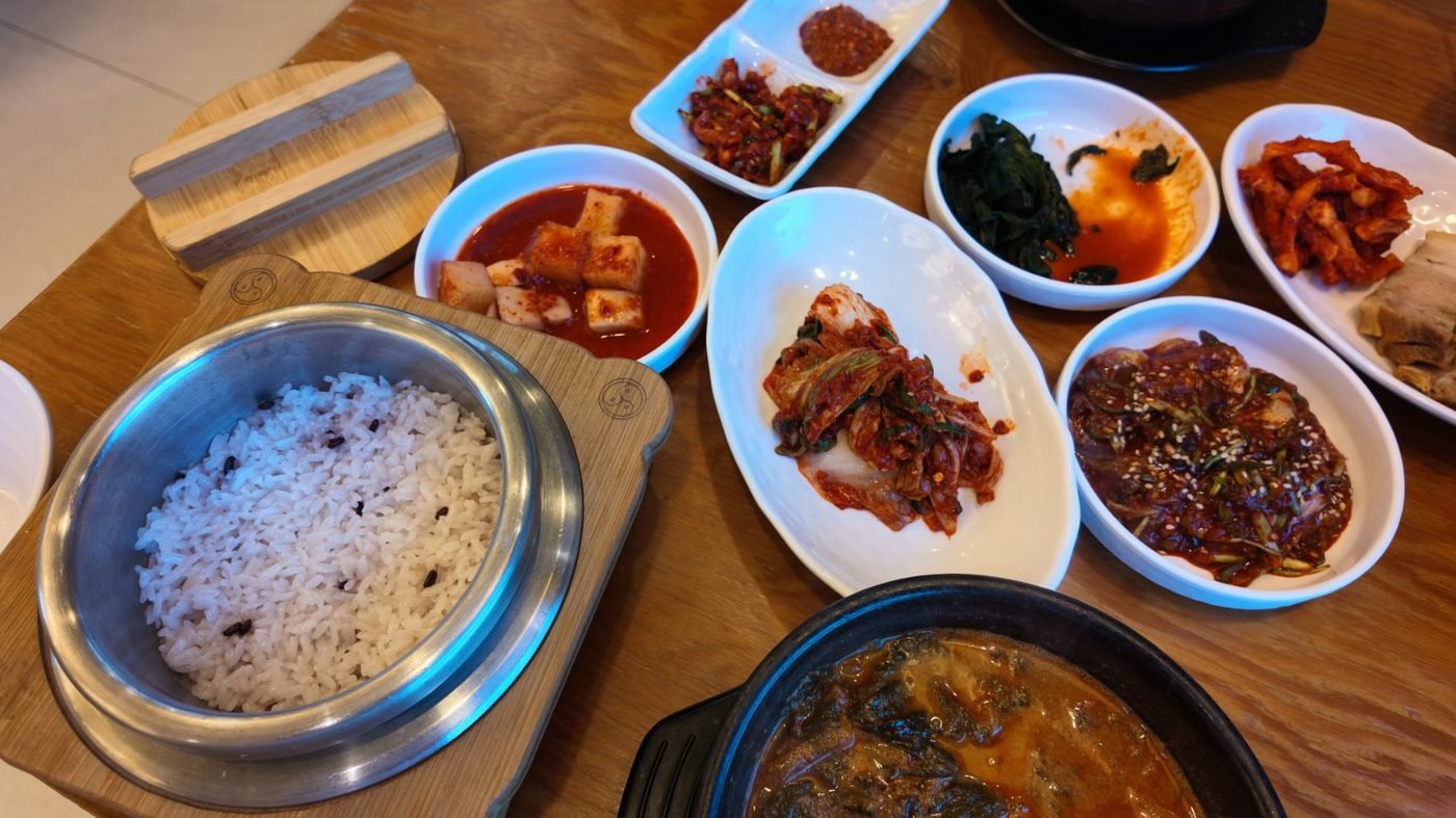 A stone pot of steaming rice flecked with black grains beside a full spread of side dishes and a dark bowl of chueotang