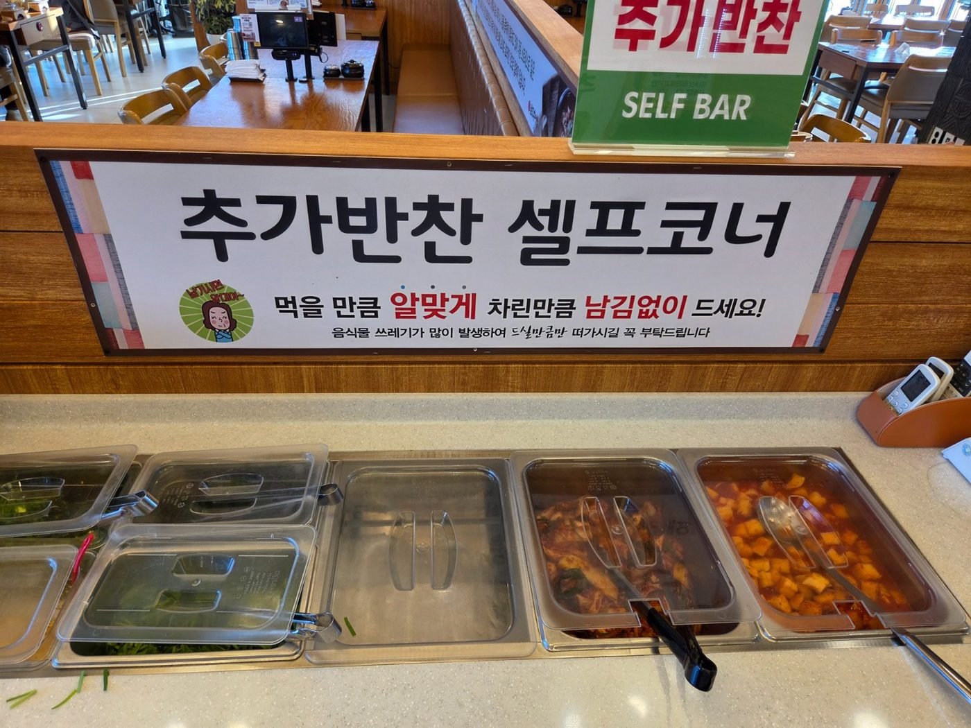 Steel trays of extra side dishes under a green sign reading 추가반찬 셀프코너, the free self-serve refill bar