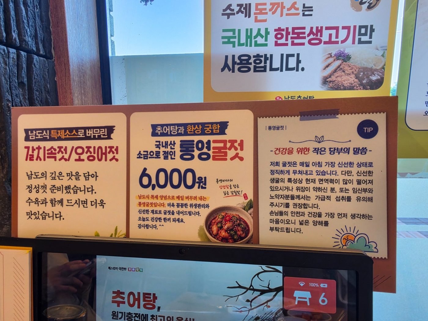 Korean signboards priced 6,000 won for Tongyeong salted oysters, beside a card describing salted hairtail and squid side dishes
