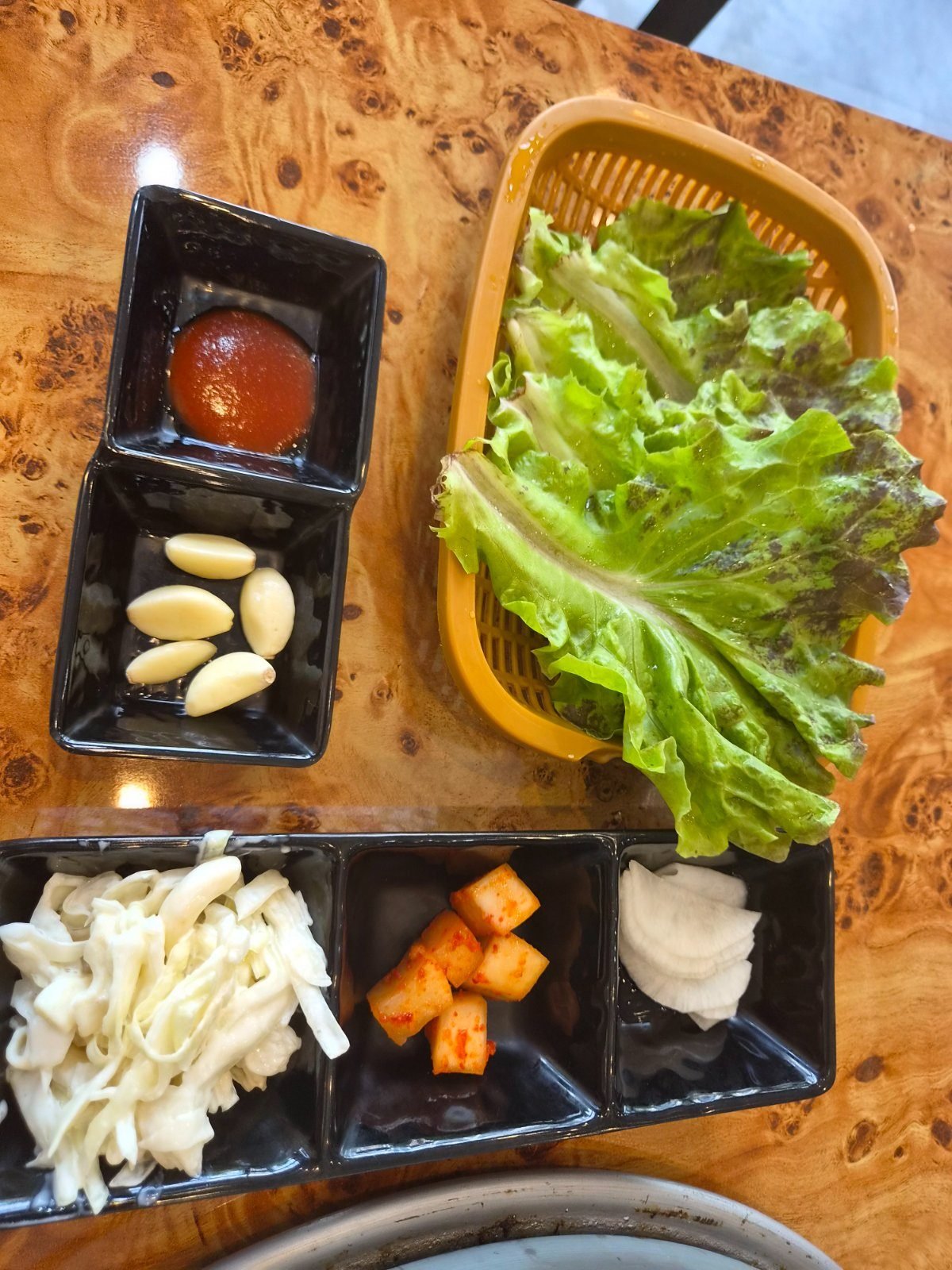 Side dishes on a wooden table: a basket of green lettuce leaves, red chojang dipping sauce, raw garlic cloves, shredded cabbage salad, cubed radish kimchi and sliced pickled radish