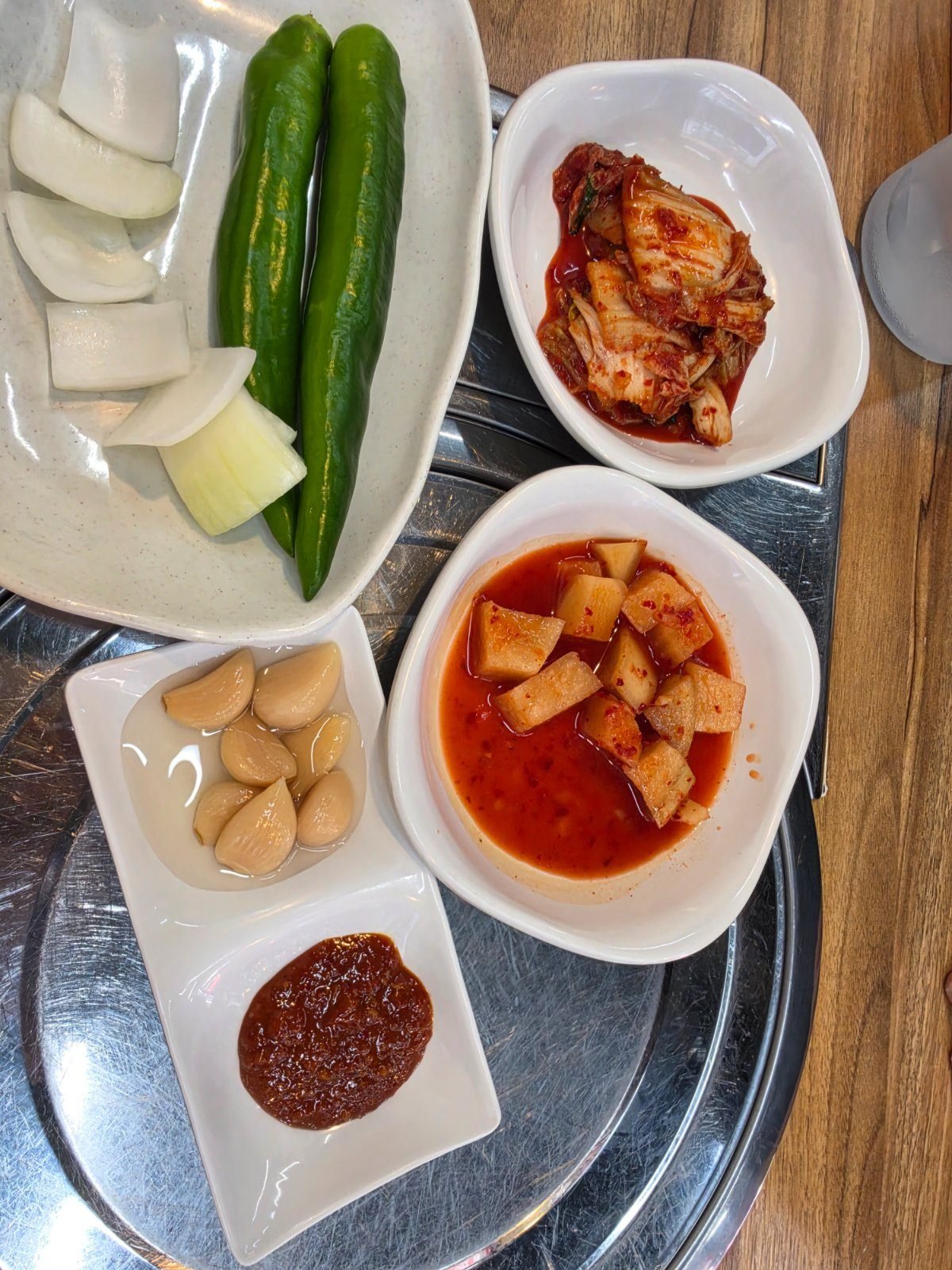 Side dishes on a steel tray: raw onion wedges, two green chillies, cabbage kimchi, cubed radish kimchi, pickled garlic cloves and ssamjang