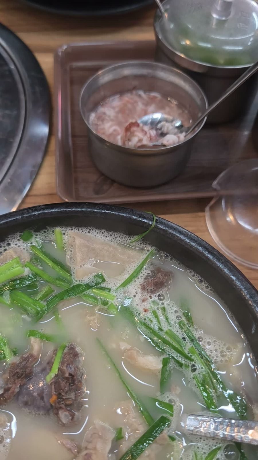 Steel pot of salted shrimp with a spoon, on a wooden caddy beside a bowl of milky pork soup