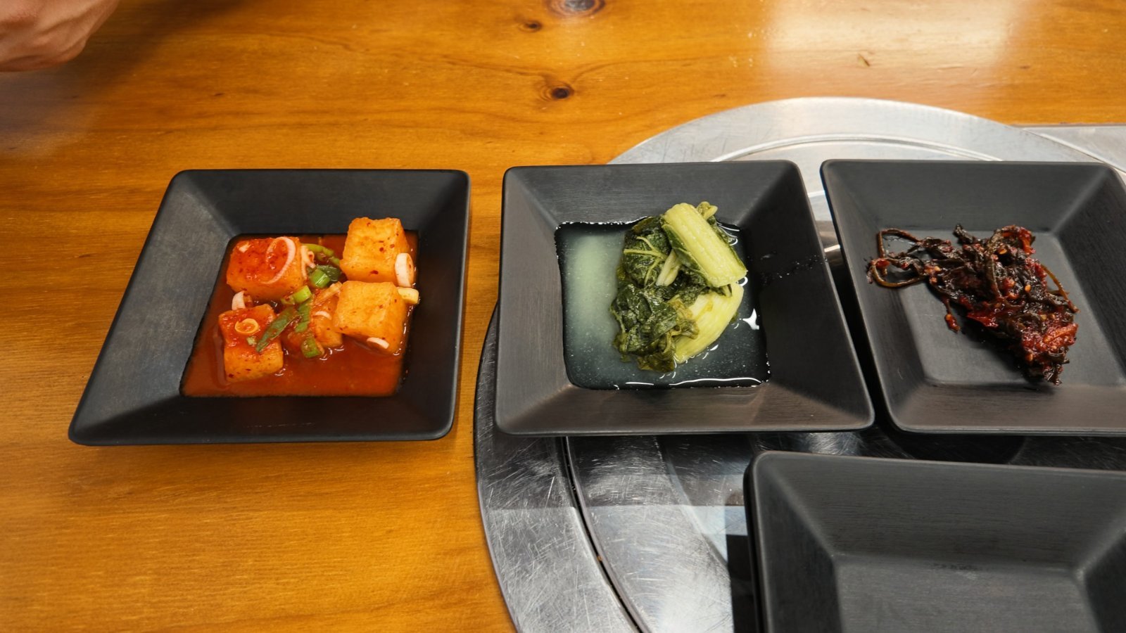 Three black square dishes on a wooden table holding red cubed radish kimchi, pale pickled greens in brine and dark salty pickles