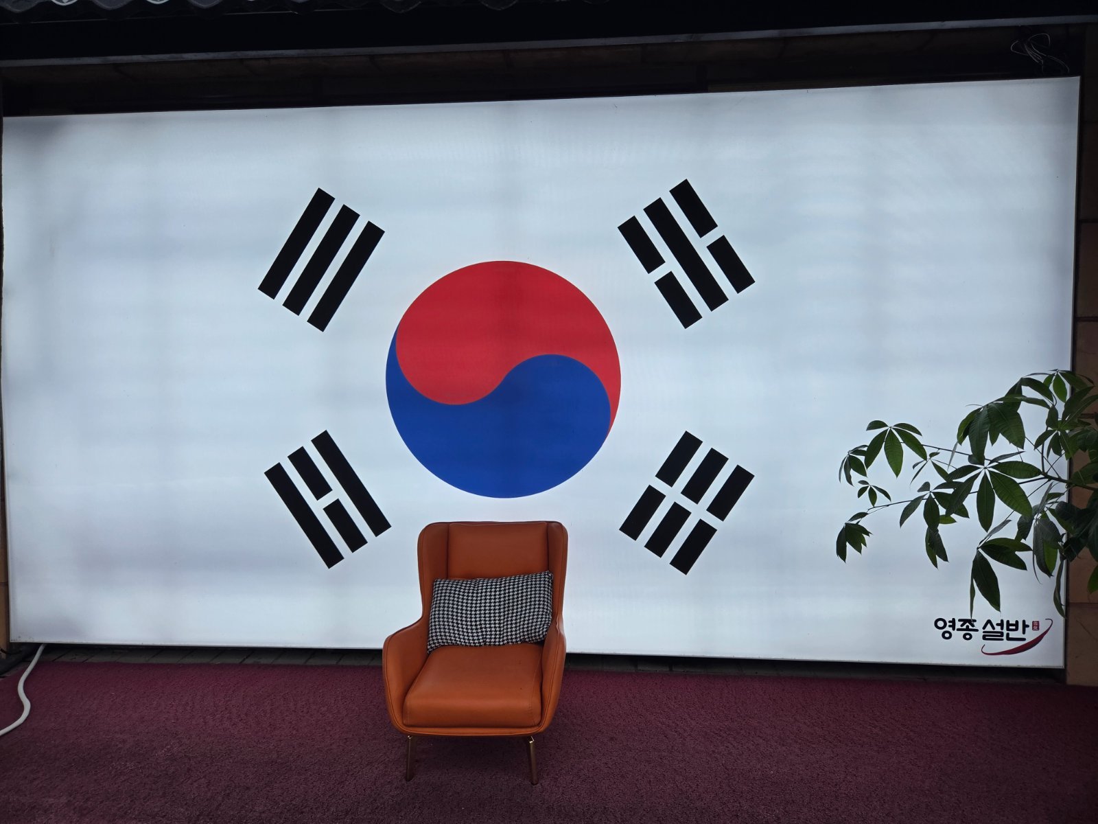 Wall-sized backlit Korean flag with a single orange leather armchair in front of it, set up as a photo corner inside the restaurant