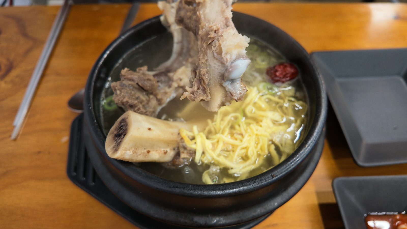 Stone pot of ugaltang, a clear beef short-rib soup with yellow egg strips, a red jujube and a cut marrow bone visible in the pale broth