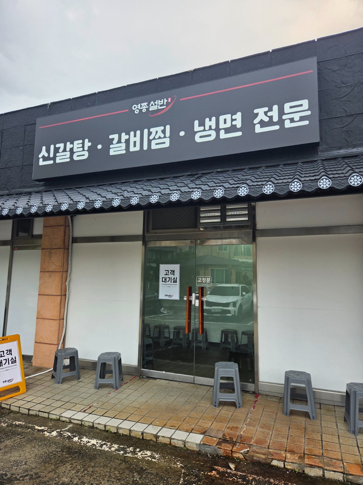 Black-tiled single-storey restaurant with a dark signboard reading 영종설반 신갈탕 갈비찜 냉면 전문, grey plastic stools lined up outside the glass doors