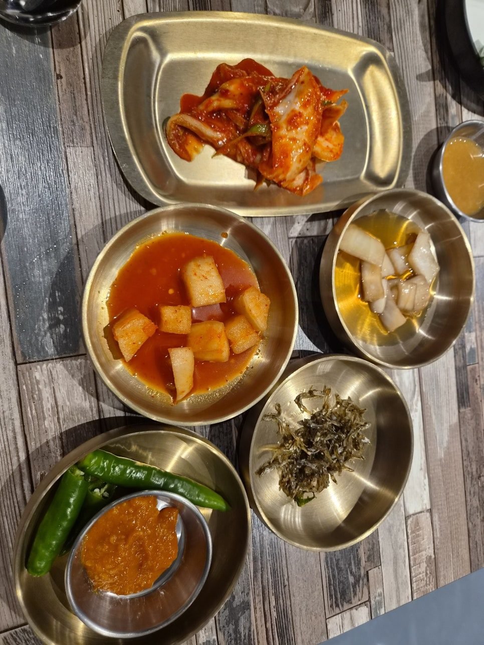 Side dishes in brass bowls: cubed radish kimchi in red brine, pickled white radish, dried anchovies, and two raw green chillies with a dish of ssamjang.