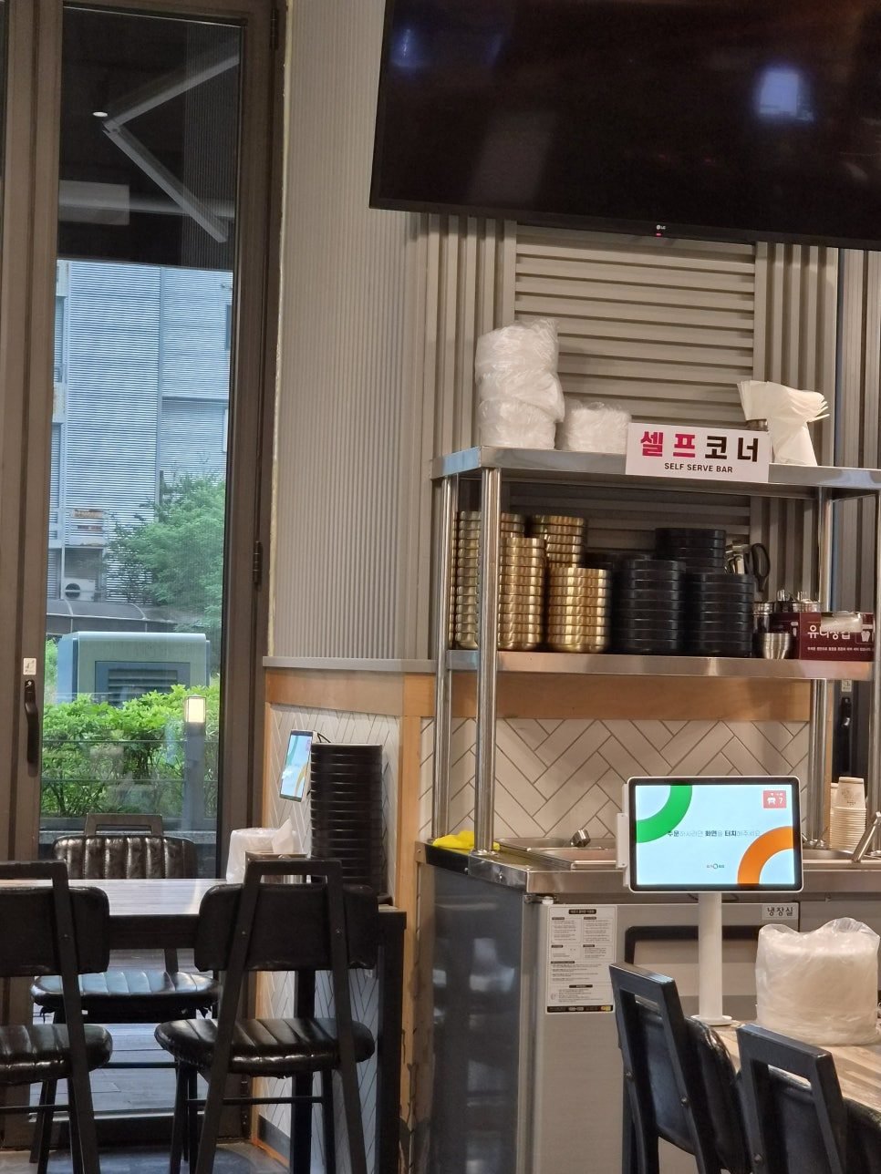 The self serve bar signed in Korean and English, with stacked brass rice bowls, black lids, water jugs and paper cups.