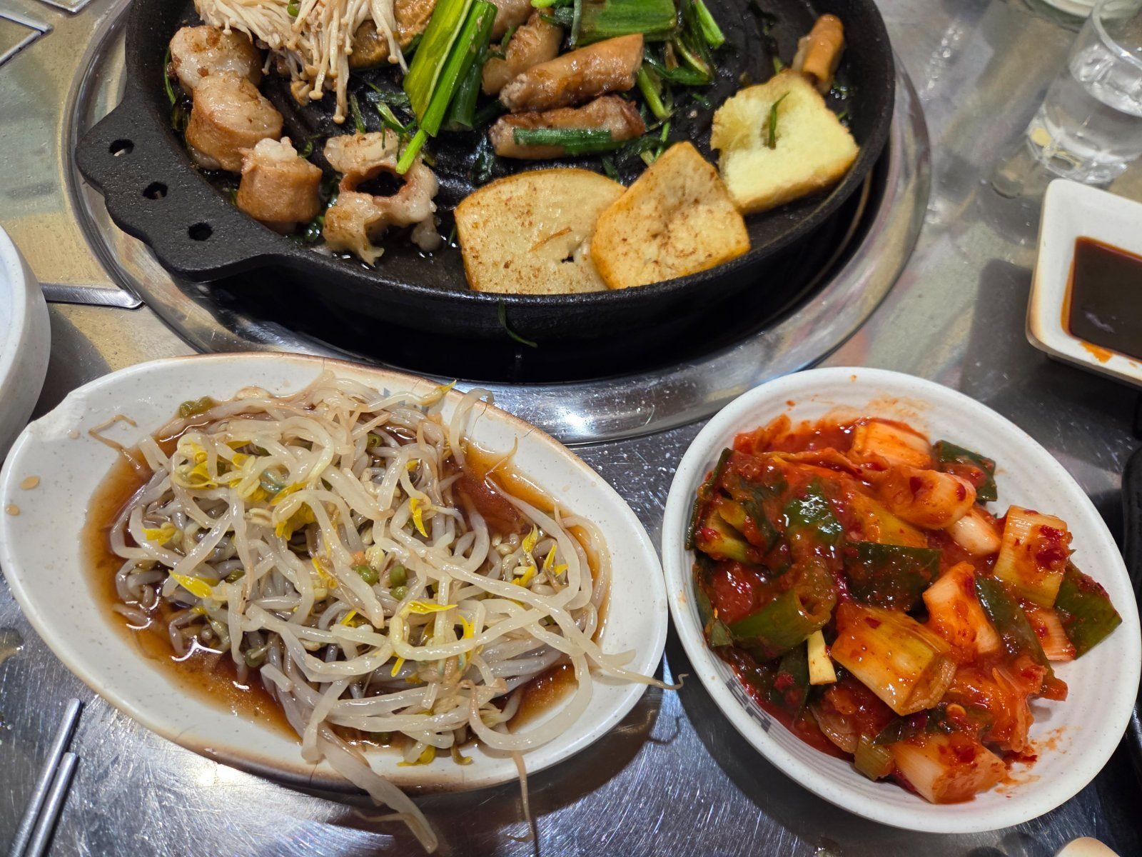 Two banchan dishes on a steel table, seasoned bean sprouts in soy dressing and chopped kimchi, next to a griddle with potato slices
