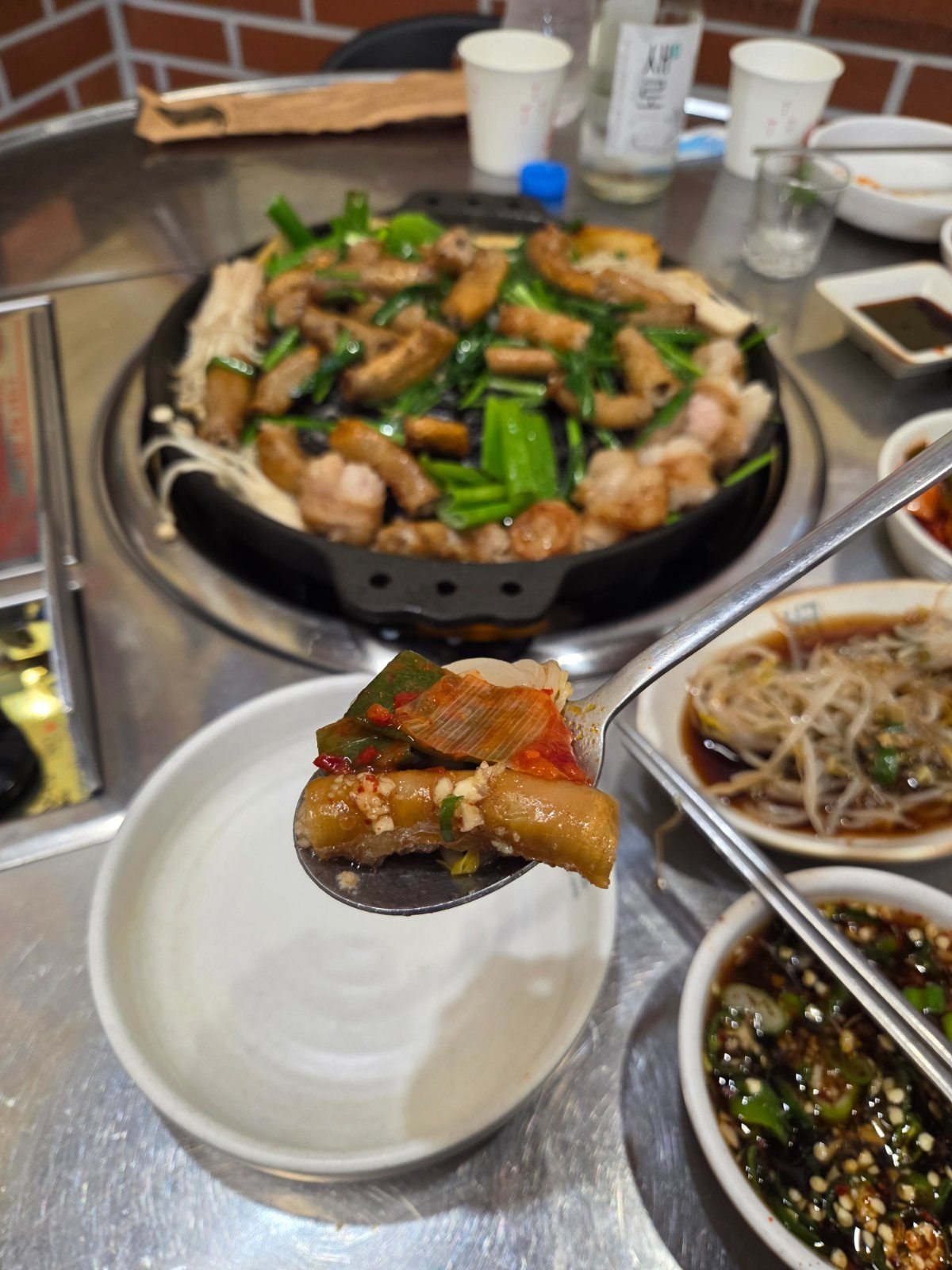 A spoon holding a browned piece of gopchang topped with red kimchi geotjeori, held over a bowl beside the griddle