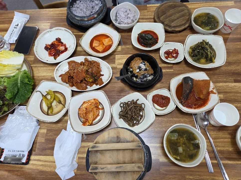 A wooden table covered in about a dozen small white dishes of banchan, a black stone pot of multigrain rice, a plate of red stir-fried pork, a black earthenware pot of ssamjang, braised fish in red sauce, kimchi and a bowl of soup.