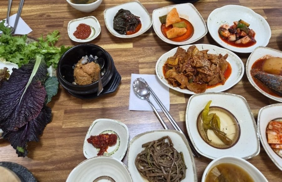 Close-up of a black earthenware pot holding a mound of brown ssamjang with pale freshwater snail meat around it, beside a pile of green lettuce and purple perilla leaves and a plate of red stir-fried pork.