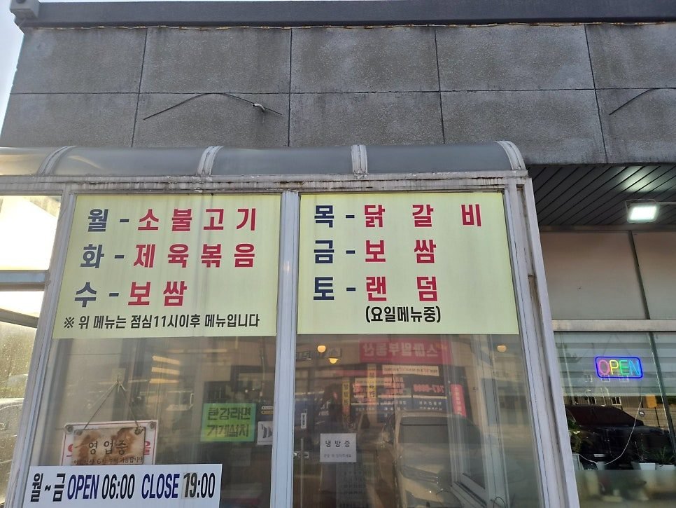 Yellow signboards listing the main dish for each weekday in Korean, with a white banner below reading Mon to Fri OPEN 06:00 CLOSE 19:00.