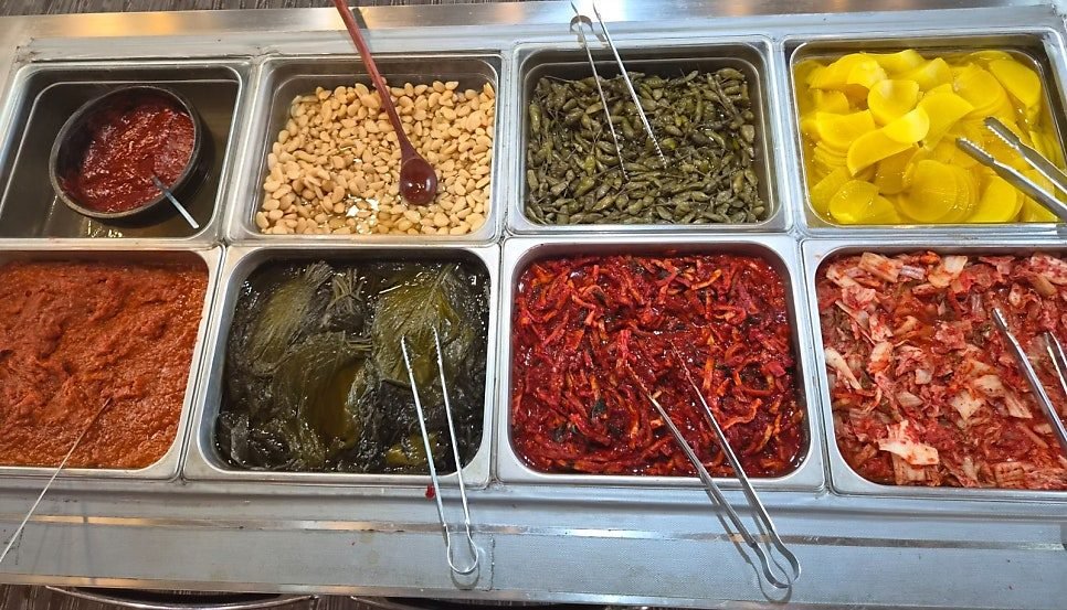 Eight stainless bins of Korean side dishes including braised soybeans, pickled green chillies, yellow pickled radish, shredded radish salad and fresh kimchi, each with its own tongs.