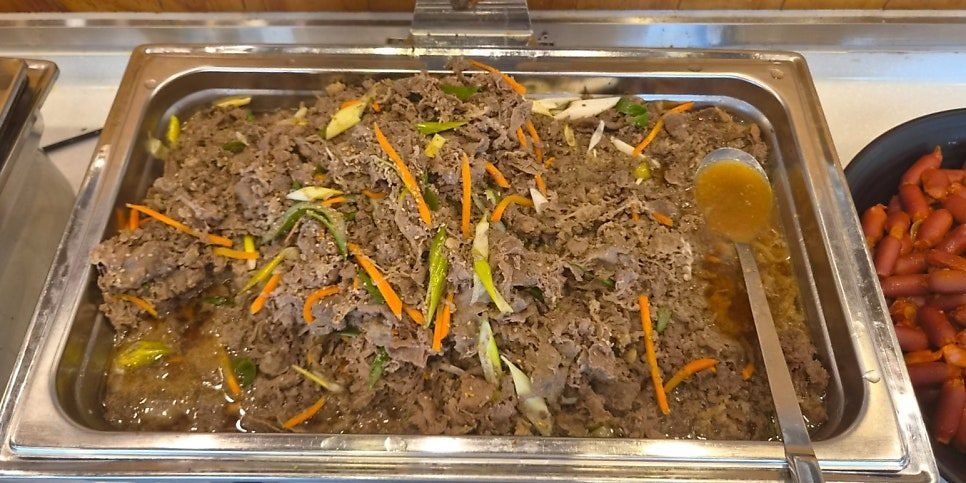 A large stainless tray of thin-sliced beef bulgogi in its own juices with carrot and onion on the heated buffet line.