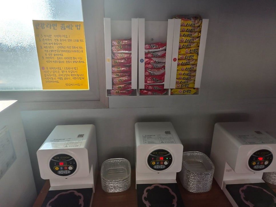 A wall rack holding three numbered slots of instant noodles - Yeol-ramyeon, Kimchi ramyeon and Snack-myeon - above the self-cook noodle machines.