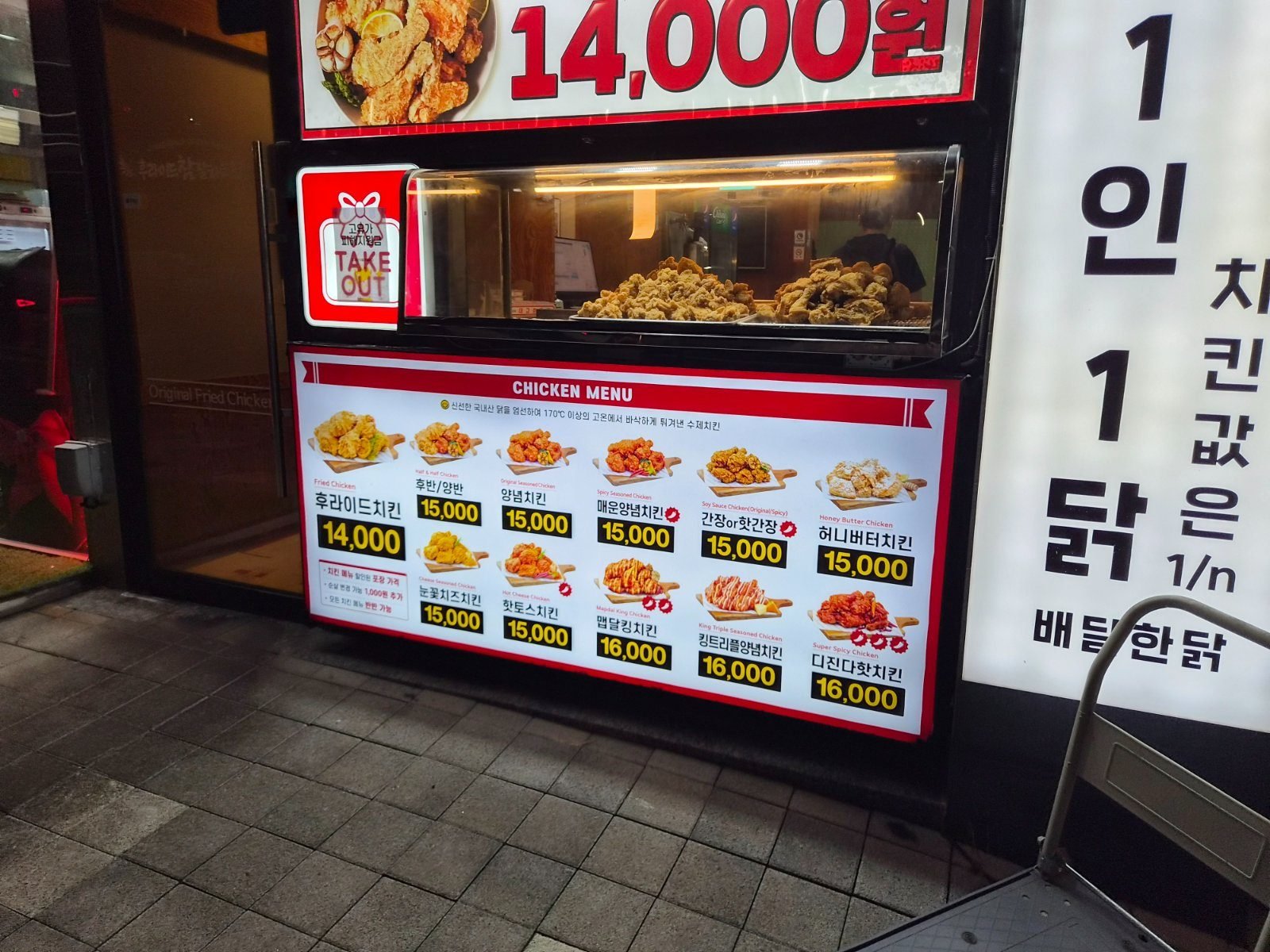 Outdoor chicken menu board with Korean and English names and prices from 14,000 to 16,000 won beside a warming case of fried chicken