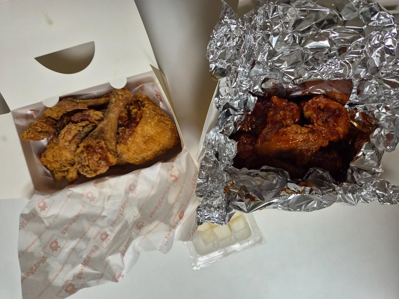 Two opened chicken portions side by side, plain fried chicken in a paper box and red seasoned chicken in foil