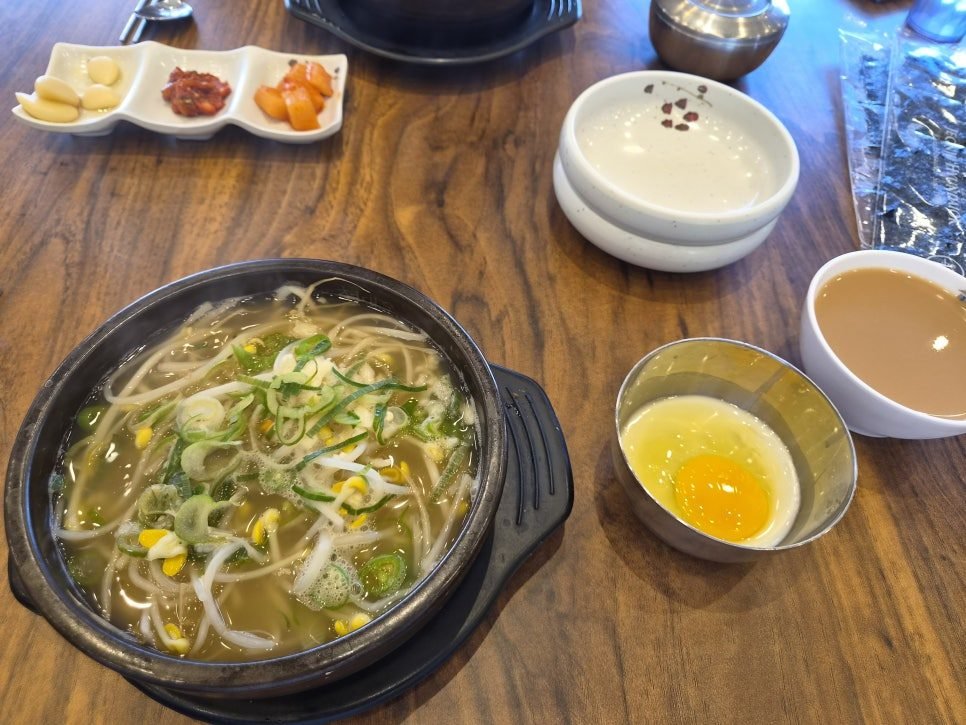 A dark stone bowl of clear bean sprout soup topped with sliced spring onion and rings of green chili, on a wooden table beside a three-part white dish of pickled garlic, red salted squid and radish kimchi, a steel bowl holding a barely-set egg, and a white cup of brown moju.