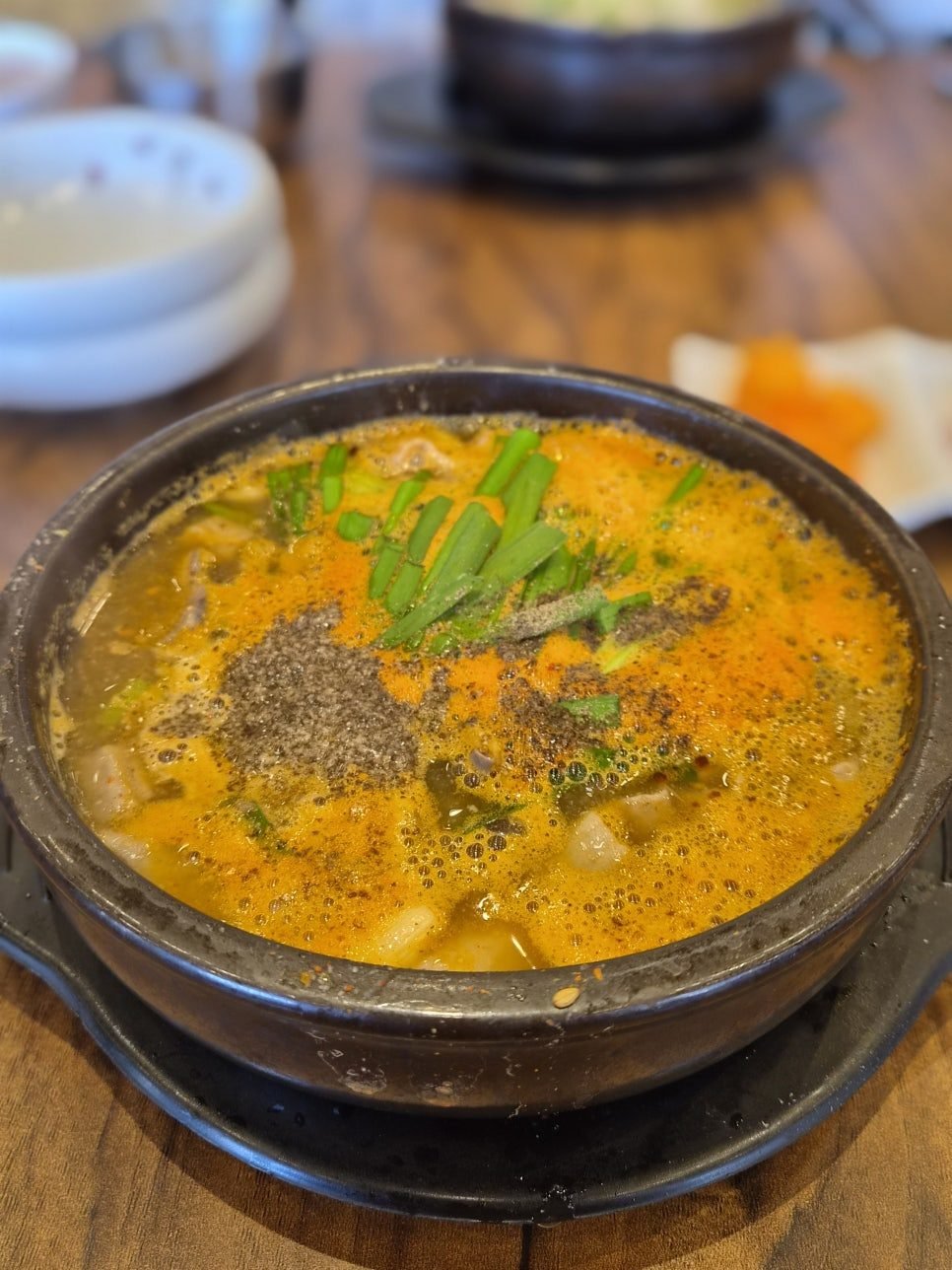 A black earthenware pot of bubbling orange-red soup on a wooden table, topped with a mound of dark perilla seed powder and cut green chives, with pale slices of pork visible in the broth.