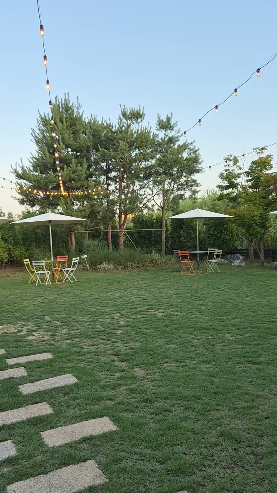 Cafe lawn at dusk with stepping stones, parasols, metal chairs and string lights in the trees