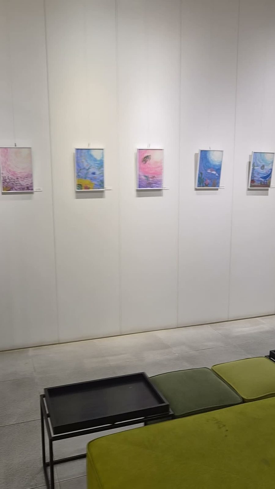 Five framed sea paintings hung on a white wall above a green bench