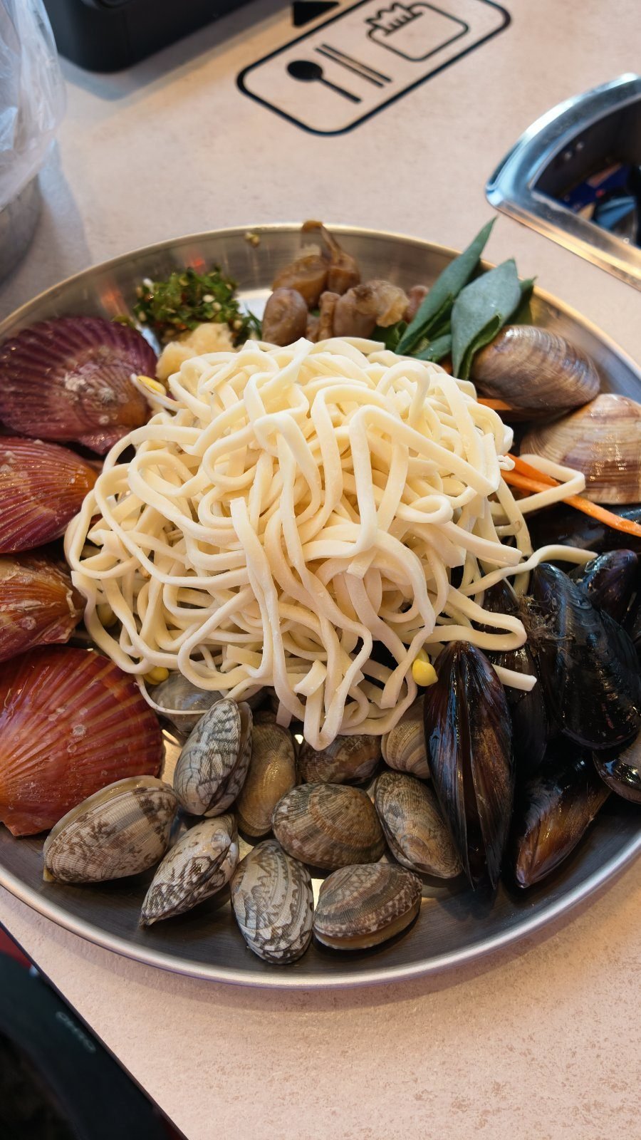 A steel platter of raw scallops, mussels and clams with a mound of fresh white kalguksu noodles piled on top, sitting on a table burner.