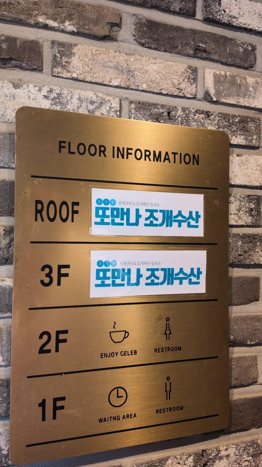 Brass floor directory plate reading FLOOR INFORMATION, with 또만나 조개수산 labels beside ROOF and 3F, and a cafe and restroom icon beside 2F.