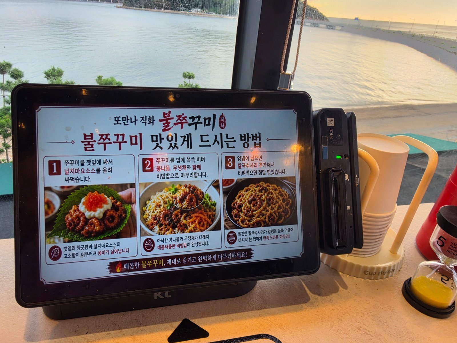 Ordering tablet on a restaurant table showing a three-step illustrated guide to eating bul-jjukkumi, with the sea and a low sun visible through the window behind it.