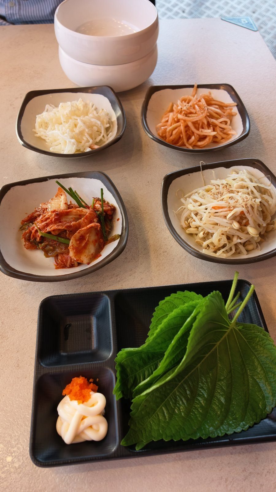 Four small black dishes of side dishes with a tray of perilla leaves and lettuce, and a white sauce topped with orange flying fish roe.