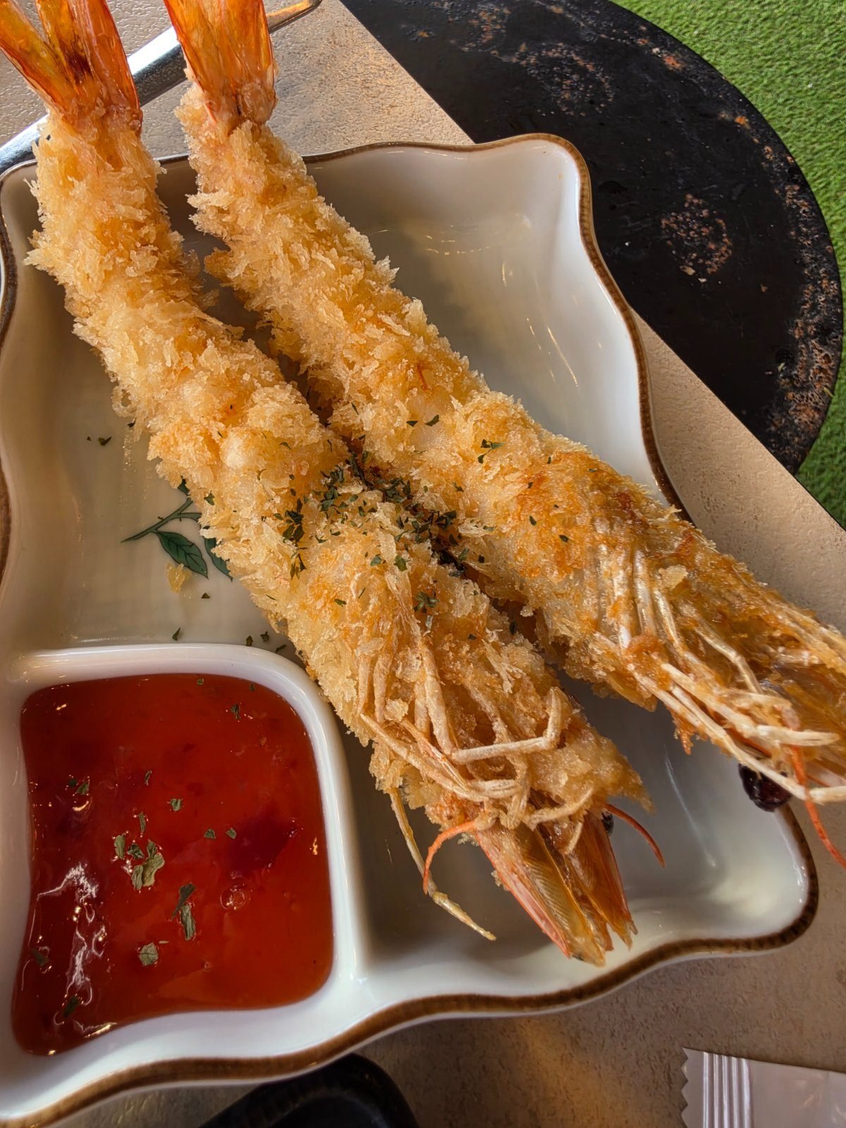 Two long breaded king prawns on a wooden board beside a dish of red dipping sauce.
